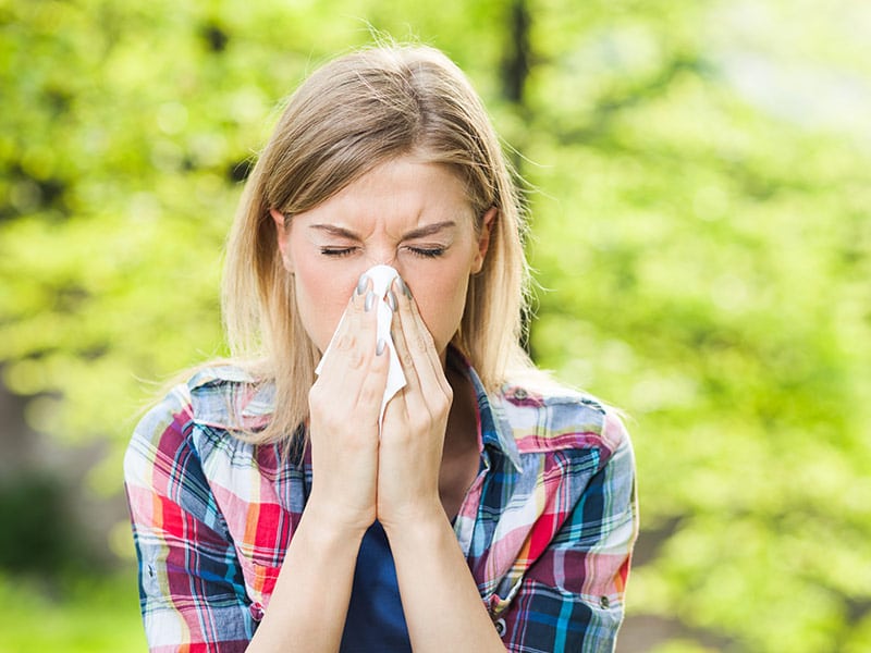 Woman with eyes closed in plaid shirt blowing her nose into a tissue standing outside with a green tree in the background.