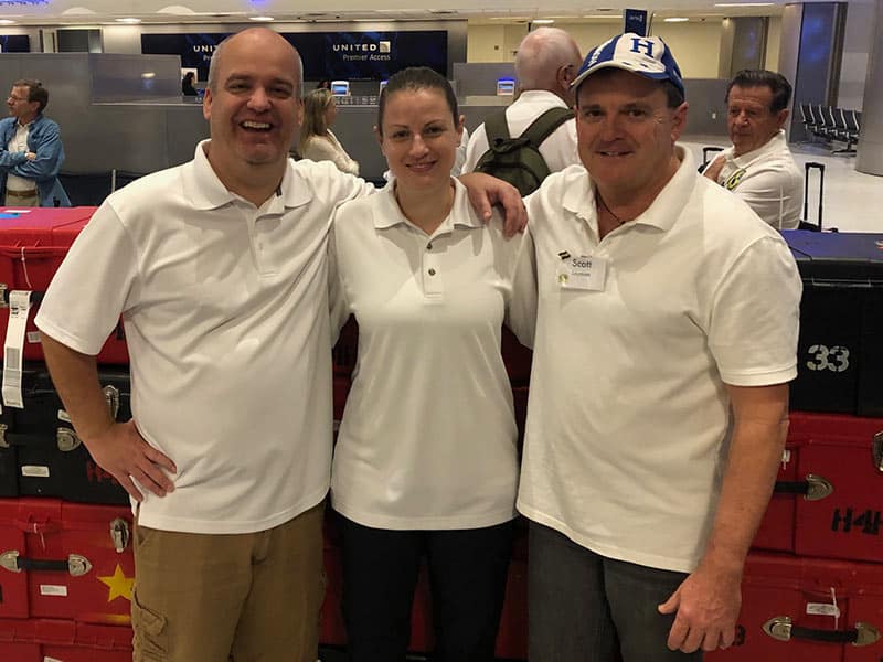 Two men and a woman in white shirts stand with arms around each other smiling at the airport in front of stacks of cases.