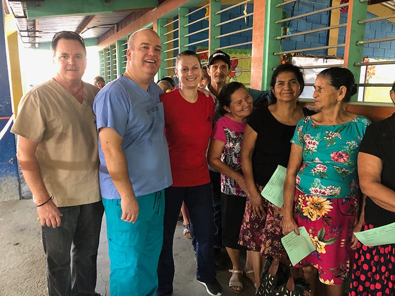 Three Methodist employees stand on a covered porch with a group of Honduran woman who are wearing bright colored clothes.