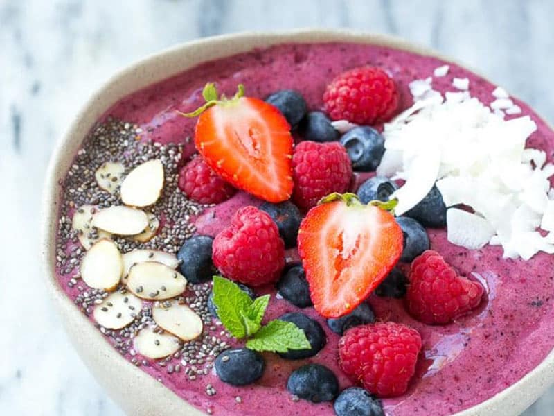 Acai bowl topped with sliced strawberries, raspberries, blueberries, sliced almonds, chia, coconut flakes and a mint leaf.