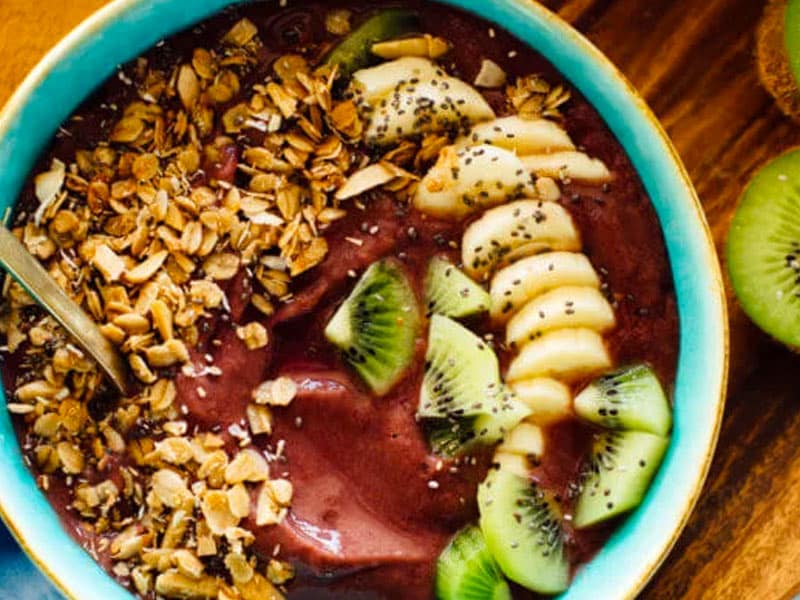 Acai bowl topped with sliced bananas and kiwi and granola in a blue bowl on a wooden countertop.