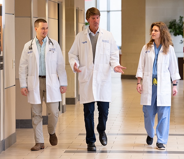 Three doctors walk down a hospital hallway wearing white lab coats. The middle doctor talks and the other two look at him.