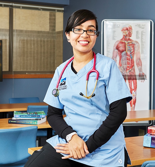 Nurse sitting on desk in scrubs with stethoscope around her neck. Poster of human muscles is in the background.