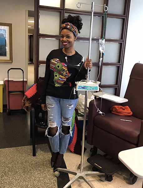 Woman standing in hospital waiting room in a sweatshirt, ripped jeans, heels and a head wrap holding an IV stand.