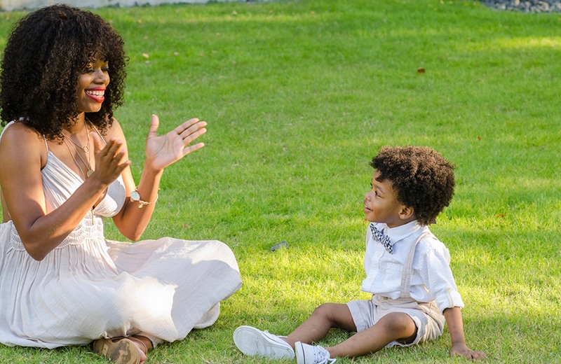 Mother sitting in grass in white dress clapping and smiling at toddler sitting next to her wearing a bow tie and suspenders.