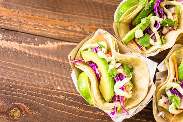 Three sets of tacos filled with avocado and cabbage and cilantro sit on a brown wooden table. 