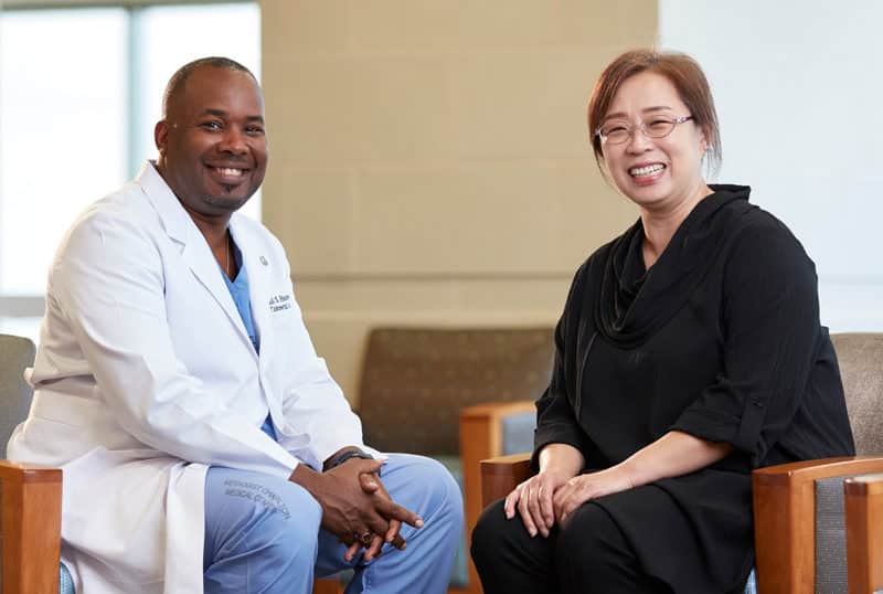 Doctor on left, sitting, wearing scrubs and patient on right, sitting, wearing black.